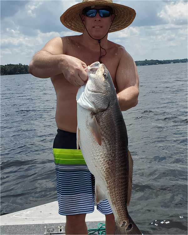 man in a bathing suit with a straw hat holding a huge redfish on the bow of a boat