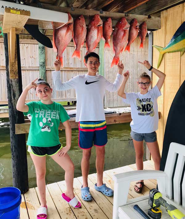 3 kids on a dock pointing to the red snapper they have hooked on the wall of a covered dock