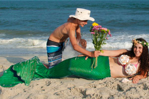 Boy kneeling in front of mermaid laying in the sand giving her flowers