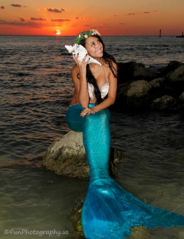 miss mermaid listening to a sea shell sitting on rocks with blazing orange sunset in background