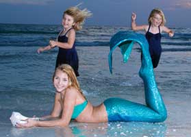 Two girls jumping around a mermaid laying in the surf