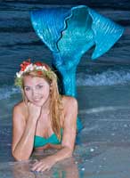 Closeup of a pretty long haired blond mermaid with little red flowers in her hair laying in the surf, holding her head with her hand resting on her elbow